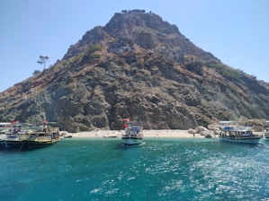 A steep, rocky mountain is surrounded by a clear blue sea, with several boats moored at a small, sandy beach. A few trees cling to the mountainside, and the contrasting colors of the sea, rock, and sky create a vibrant scene.