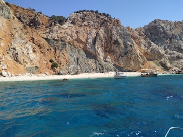 A pristine beach bordered by dramatic cliffs with rugged, earthy tones. The crystal-clear blue water gently laps at the sandy shore. Two boats are anchored near the beach, adding a touch of serenity to the natural landscape. Sparse greenery dots the rocky cliffs.