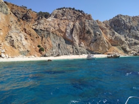 A pristine beach bordered by dramatic cliffs with rugged, earthy tones. The crystal-clear blue water gently laps at the sandy shore. Two boats are anchored near the beach, adding a touch of serenity to the natural landscape. Sparse greenery dots the rocky cliffs.