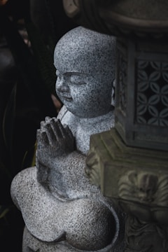 A stone sculpture of a child monk in a praying position, with hands clasped together, set against a backdrop of dark foliage. The sculpture has smooth, rounded features and appears to be part of a serene garden setting.