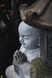 A stone statue of a child monk with hands clasped in prayer. The statue exhibits a detailed, speckled texture and is partially sheltered by an ornate stone structure.