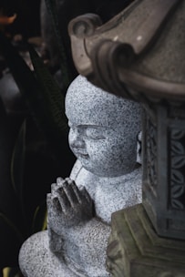 A stone statue of a child monk with hands clasped in prayer. The statue exhibits a detailed, speckled texture and is partially sheltered by an ornate stone structure.