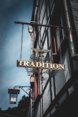 A vintage metal sign with ornate details featuring the word 'Tradition' hangs from the side of a half-timbered building. The sign is adorned with a decorative eagle sculpture on top. In the background, the building's architecture is characterized by timber framing and red window accents. A traditional street lantern is mounted near the sign.