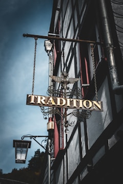 A vintage metal sign with ornate details featuring the word 'Tradition' hangs from the side of a half-timbered building. The sign is adorned with a decorative eagle sculpture on top. In the background, the building's architecture is characterized by timber framing and red window accents. A traditional street lantern is mounted near the sign.