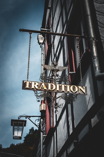 A vintage metal sign with ornate details featuring the word 'Tradition' hangs from the side of a half-timbered building. The sign is adorned with a decorative eagle sculpture on top. In the background, the building's architecture is characterized by timber framing and red window accents. A traditional street lantern is mounted near the sign.