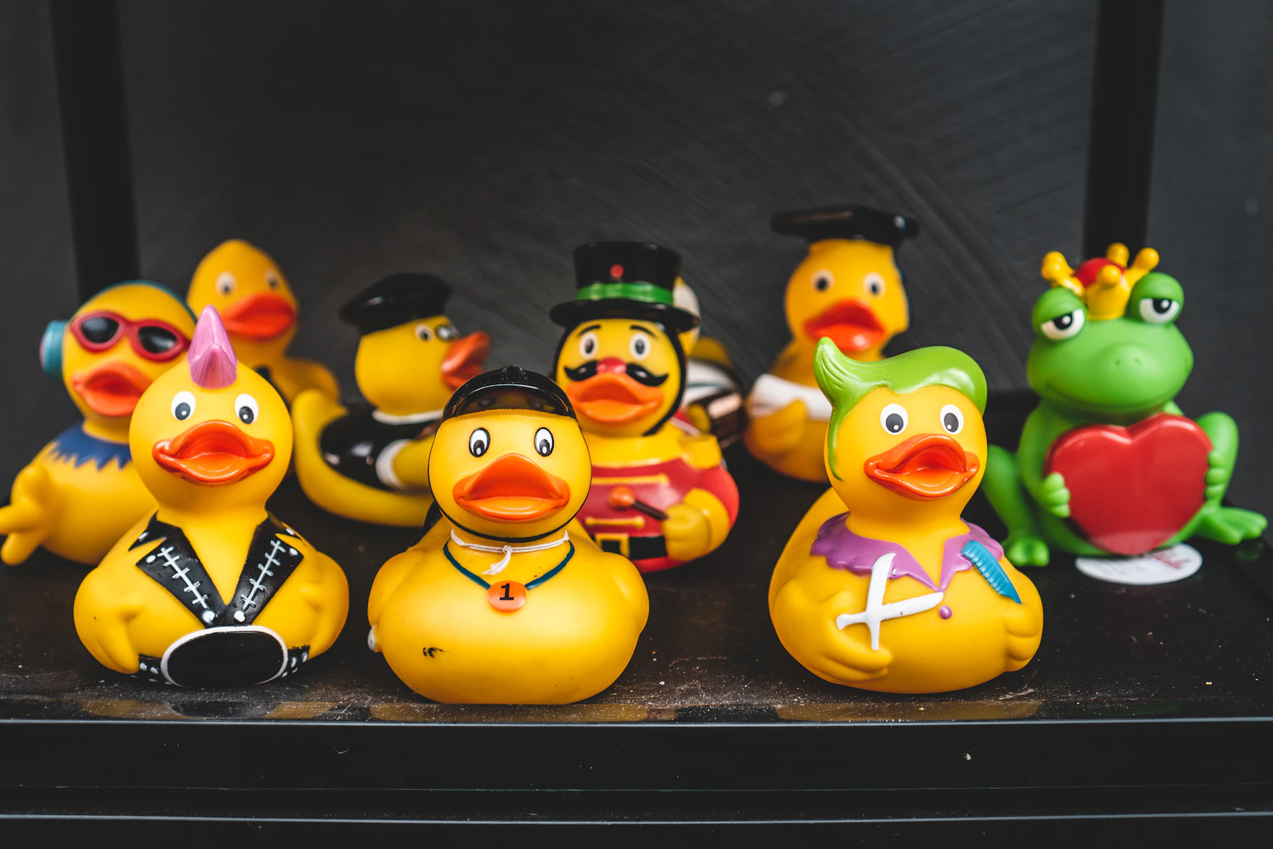 Rubber ducks in costumes, with one frog among them, lined up on a dark shelf