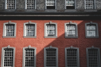 A red-brick building facade featuring multiple windows with decorative grey trim and architectural accents. The building has a symmetrical arrangement of windows in rows, capturing a mix of shadow and sunlight on the brick surface.