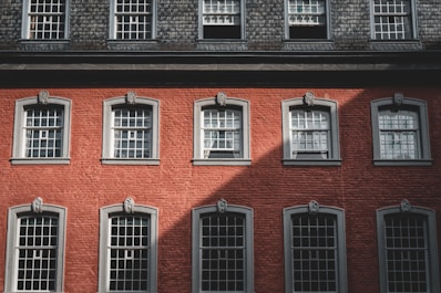 A red-brick building facade featuring multiple windows with decorative grey trim and architectural accents. The building has a symmetrical arrangement of windows in rows, capturing a mix of shadow and sunlight on the brick surface.