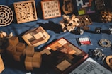 A collection of various wooden and metal puzzles is displayed on a dark fabric background. The assortment includes intricate 3D structures, interlocking block puzzles, number puzzles, and labyrinth games. The puzzles feature a variety of shapes and sizes, with some laid out flat and others standing upright, casting shadows in the light.
