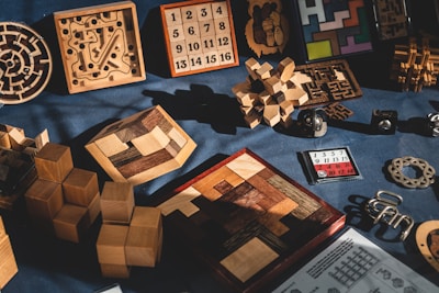 A collection of various wooden and metal puzzles is displayed on a dark fabric background. The assortment includes intricate 3D structures, interlocking block puzzles, number puzzles, and labyrinth games. The puzzles feature a variety of shapes and sizes, with some laid out flat and others standing upright, casting shadows in the light.