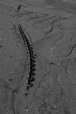 A long, heavy chain partially buried in the sand on a beach. The chain appears to extend into the distance, with visible links showing signs of rust and weathering. The sand is textured with lines and small scattered pebbles.