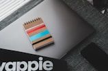 A spiral notebook with a cardboard cover adorned with colorful stripes in pink, red, blue, and black rests on a closed laptop with a sleek design. A dark-textured fabric surface supports these items, adding a subtle contrast. Nearby, part of a smartphone and a partial view of a black object with the lettering stylized possibly from a brand or a product are visible.