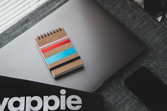A spiral notebook with a cardboard cover adorned with colorful stripes in pink, red, blue, and black rests on a closed laptop with a sleek design. A dark-textured fabric surface supports these items, adding a subtle contrast. Nearby, part of a smartphone and a partial view of a black object with the lettering stylized possibly from a brand or a product are visible.