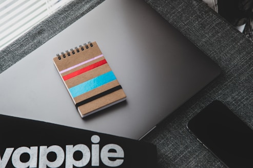 A spiral notebook with a cardboard cover adorned with colorful stripes in pink, red, blue, and black rests on a closed laptop with a sleek design. A dark-textured fabric surface supports these items, adding a subtle contrast. Nearby, part of a smartphone and a partial view of a black object with the lettering stylized possibly from a brand or a product are visible.