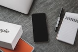 A neatly arranged flat lay on a textured grey surface features a smartphone, a closed laptop, a pen, and a spiral notepad. Additionally, there is a branded product box partially opened, showing an orange interior.