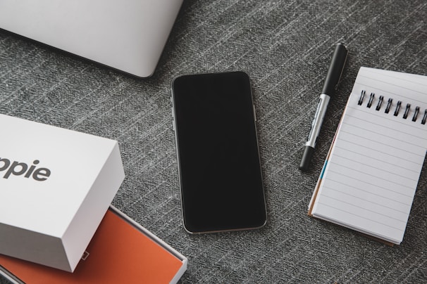 A neatly arranged flat lay on a textured grey surface features a smartphone, a closed laptop, a pen, and a spiral notepad. Additionally, there is a branded product box partially opened, showing an orange interior.