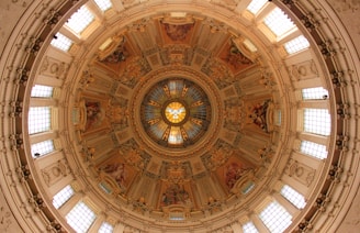 An intricately designed dome featuring detailed artwork and ornate architectural elements. The central part of the dome contains stained glass with a symmetrical design, and the surrounding areas are adorned with paintings and sculptural details.