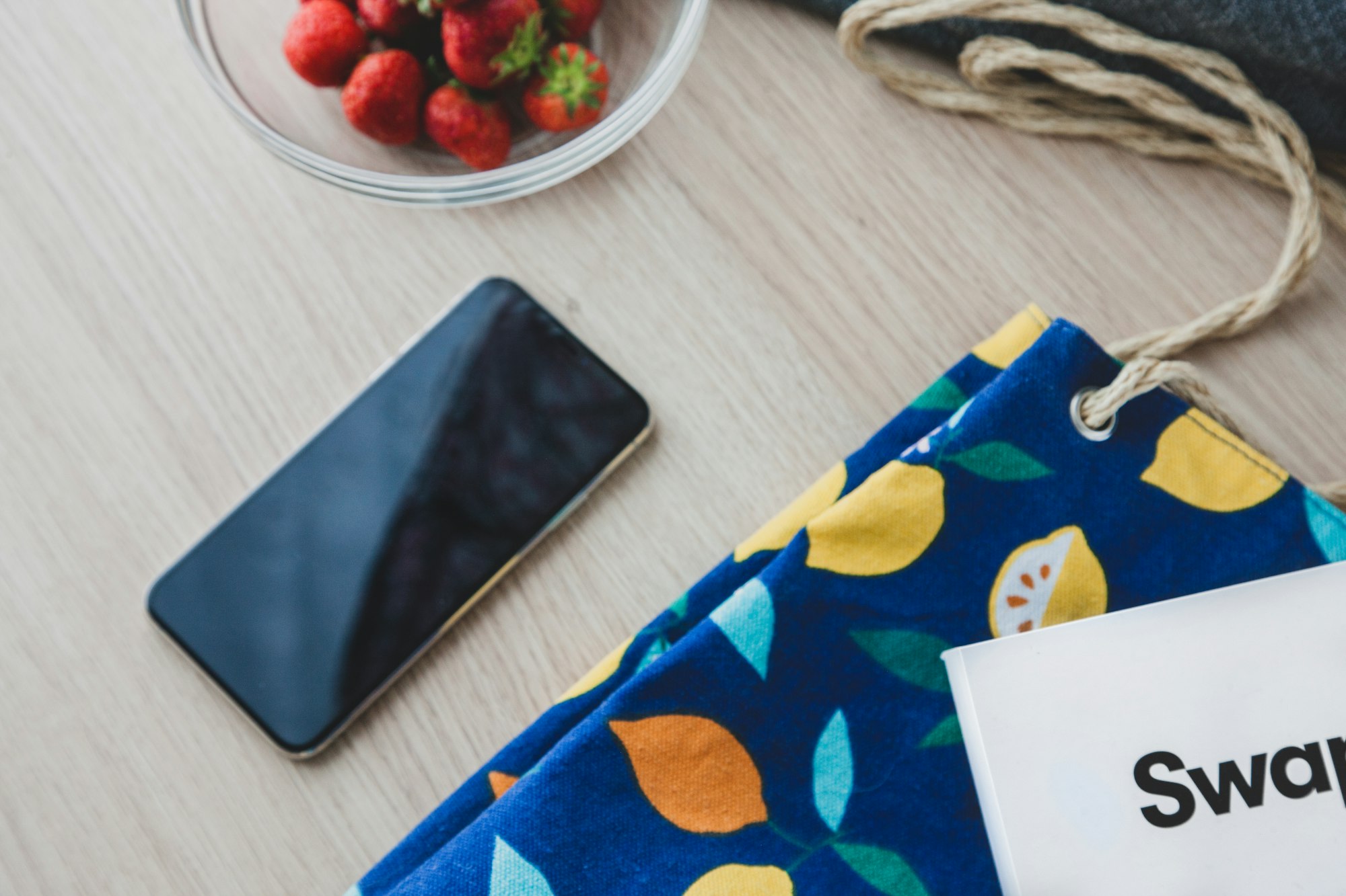 Smartphone on a breakfast table. Background is light wood. Strawberries strike with their red color. Lemons on the texture of a bag. Cozy mood.