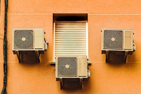 Three air conditioning units mounted on the exterior wall of a building, with a closed vent in the center. The wall is a warm, peach color and the units have a beige finish. A black cable runs vertically along the left side of the image.