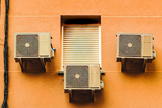 Three air conditioning units mounted on the exterior wall of a building, with a closed vent in the center. The wall is a warm, peach color and the units have a beige finish. A black cable runs vertically along the left side of the image.