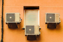 Three air conditioning units mounted on the exterior wall of a building, with a closed vent in the center. The wall is a warm, peach color and the units have a beige finish. A black cable runs vertically along the left side of the image.