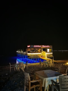 A beachside café with wooden tables and chairs is set up on the sand. A small pier extends over the water, with LED lighting illuminating the area in a soft blue glow. The café sign is lit in red, displaying 'Lider Beach'. The scene is captured at night, creating a tranquil atmosphere.