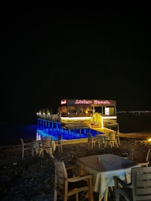 A beachside caf&eacute; with wooden tables and chairs is set up on the sand. A small pier extends over the water, with LED lighting illuminating the area in a soft blue glow. The caf&eacute; sign is lit in red, displaying 'Lider Beach'. The scene is captured at night, creating a tranquil atmosphere.