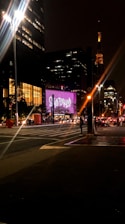 A nighttime urban scene featuring well-lit buildings and a street intersection. Bright lights are shining from street lamps and the windows of a tall building. A prominent illuminated sign displays the text 'SÃO PAULO' in a bold, pink font. A few cars and people are visible, with an illuminated tower in the distance adding to the cityscape.