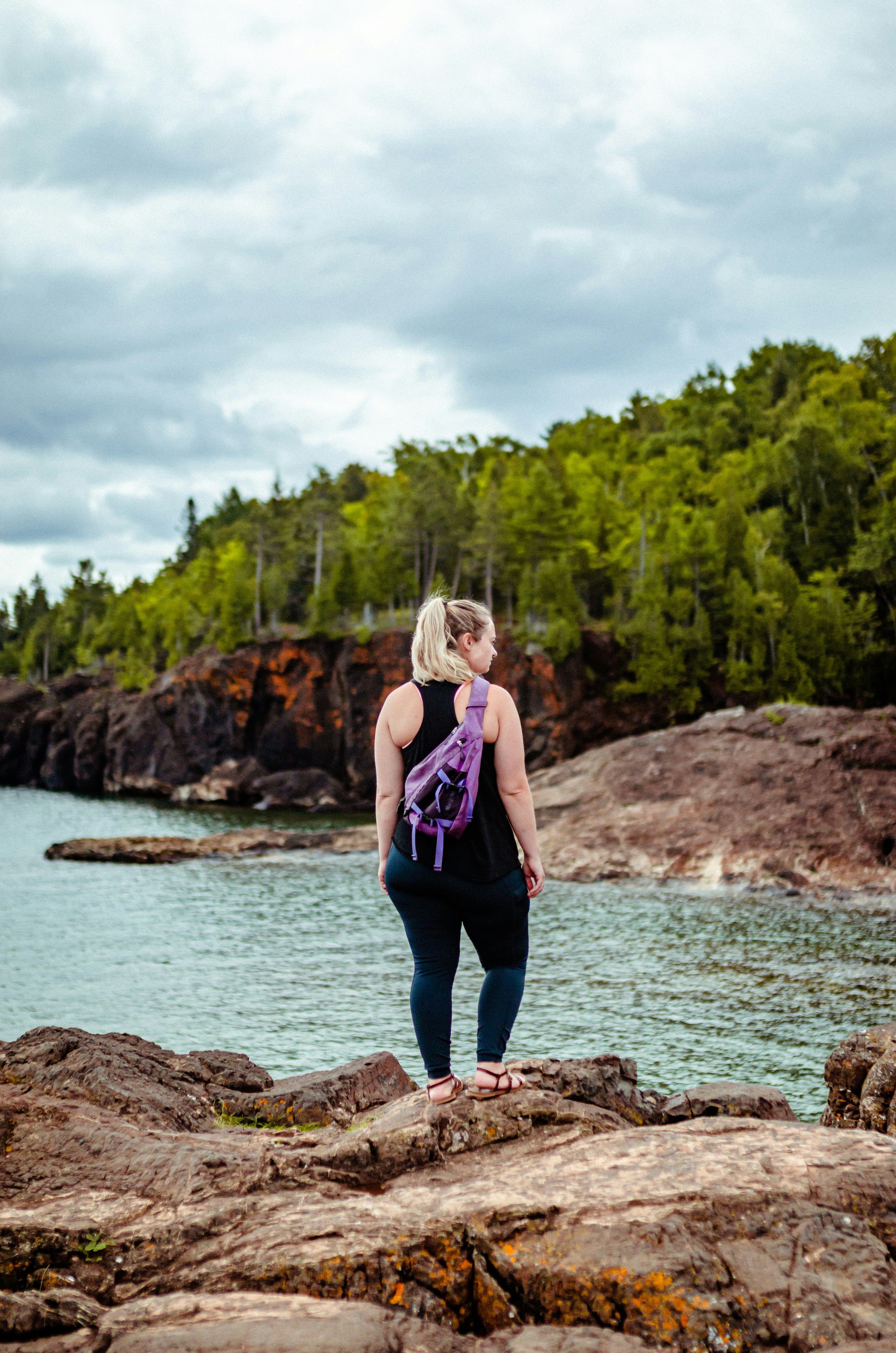 woman in purple tank top and blue denim jeans standing on rock near body of water