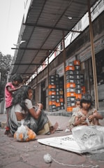 Several children and a woman are seated on a sidewalk near a building, working with marbles and string. The environment appears urban and the group seems to be engaged in a craft or task. Behind them, there are stacks of boxes organized outside a store.