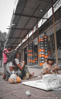 Several children and a woman are seated on a sidewalk near a building, working with marbles and string. The environment appears urban and the group seems to be engaged in a craft or task. Behind them, there are stacks of boxes organized outside a store.