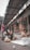 Several children and a woman are seated on a sidewalk near a building, working with marbles and string. The environment appears urban and the group seems to be engaged in a craft or task. Behind them, there are stacks of boxes organized outside a store.