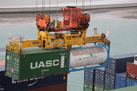 A crane mechanism is lifting a large shipping container from a stack of other containers at a port. The container has the logo 'UASC' on it and next to it is a cylinder-shaped tank marked with 'SINOCHEM LANTIAN'. The crane is painted bright yellow and orange, and the ocean and a sandy shore are visible in the background.