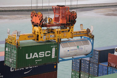 A crane mechanism is lifting a large shipping container from a stack of other containers at a port. The container has the logo 'UASC' on it and next to it is a cylinder-shaped tank marked with 'SINOCHEM LANTIAN'. The crane is painted bright yellow and orange, and the ocean and a sandy shore are visible in the background.