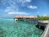 Several thatched-roof bungalows are situated over clear, turquoise water, connected by a wooden walkway. A small set of stairs leads down to the water, and lush green plants adorn the walkway. The sky is clear and bright, with a few scattered clouds.