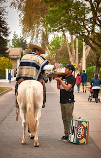A person wearing traditional attire is on horseback and is interacting with a street vendor. The vendor is holding a tray with colorful sweets and standing next to a couple of cardboard boxes. Surrounding them are trees and a few people walking further down the street.