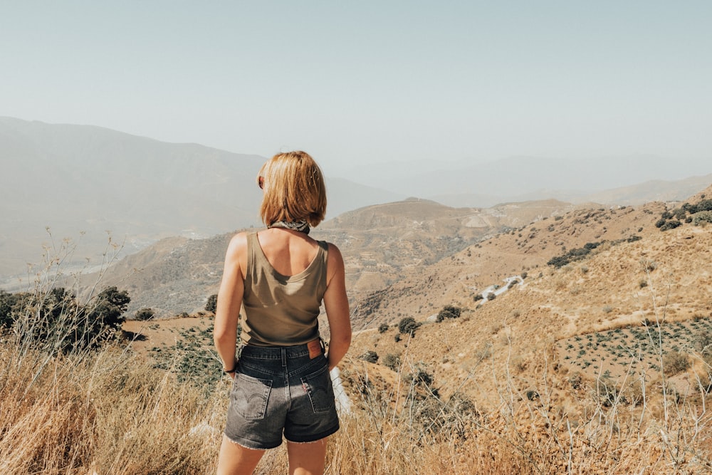 woman in brown tank top and blue denim shorts standing on brown grass field during daytime