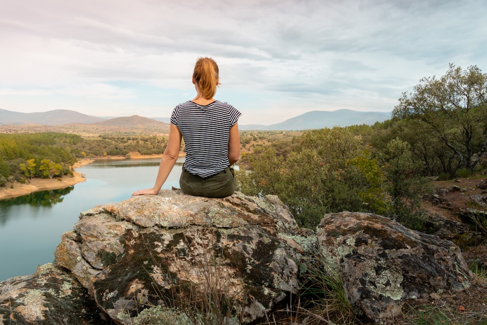 woman in black and white stripe shirt sitting on rock near lake during daytime