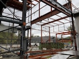 A construction site with a partially built metal framework structure. The scene is cluttered with construction materials and debris. Green containers and industrial equipment are visible in the background, with trees beyond a fence.