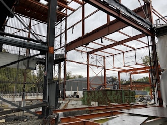 A construction site with a partially built metal framework structure. The scene is cluttered with construction materials and debris. Green containers and industrial equipment are visible in the background, with trees beyond a fence.
