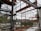 A construction site with a partially built metal framework structure. The scene is cluttered with construction materials and debris. Green containers and industrial equipment are visible in the background, with trees beyond a fence.