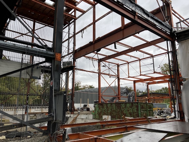 A construction site with a partially built metal framework structure. The scene is cluttered with construction materials and debris. Green containers and industrial equipment are visible in the background, with trees beyond a fence.