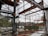 A construction site with a partially built metal framework structure. The scene is cluttered with construction materials and debris. Green containers and industrial equipment are visible in the background, with trees beyond a fence.