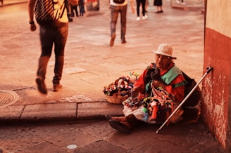 An elderly street vendor wearing a white hat sits on the pavement near a wall, surrounded by colorful handmade dolls displayed in a basket. Sunlight casts a warm glow over the scene, as pedestrians walk by in a busy, cobblestone street setting.