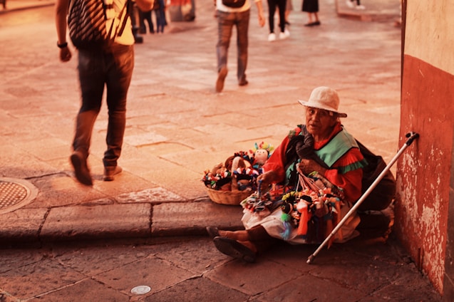 An elderly street vendor wearing a white hat sits on the pavement near a wall, surrounded by colorful handmade dolls displayed in a basket. Sunlight casts a warm glow over the scene, as pedestrians walk by in a busy, cobblestone street setting.