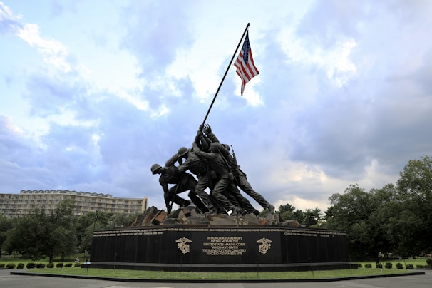 A large bronze memorial sculpture depicts soldiers raising a flag on a rugged landscape. The American flag is prominently displayed reaching up into the sky. The monument is surrounded by greenery and a cloudy sky, with buildings visible in the background.