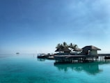 Several thatched-roof huts are built on stilts above a calm, clear turquoise sea, with palm trees around. A small boat is moored to the dock. The sky is bright blue and clear, with a distant island barely visible on the horizon.
