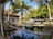 A tropical setting with palm trees surrounding a serene outdoor area. A wooden walkway crosses a reflective water feature, leading to an open, thatched-roof building with shaded seating. Lush green foliage creates a relaxing atmosphere in the bright sunlight.