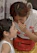 A woman is leaning close to a young girl, whispering something into her ear. The young girl listens attentively with a slight smile. The woman is wearing a white shirt with red lettering and has a red sling bag. The girl has a white sleeveless top and is wearing a necklace.