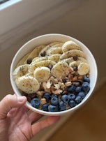 A hand holds a white bowl filled with a healthy assortment of sliced bananas, fresh blueberries, chia seeds, chocolate chips, chopped almonds, and walnuts.